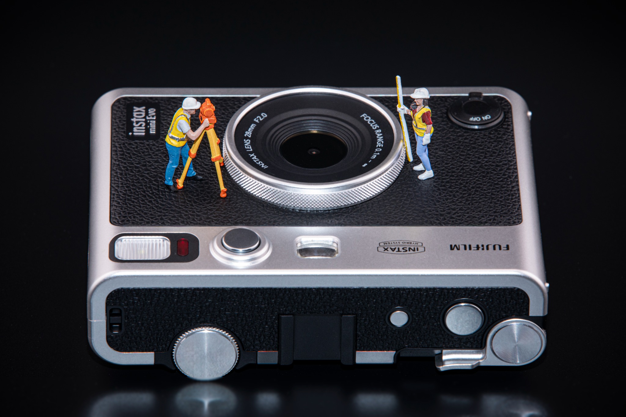 A Fujifilm INSTAX mini Evo camera photographed with two miniature surveyors beside its lens against a black background.